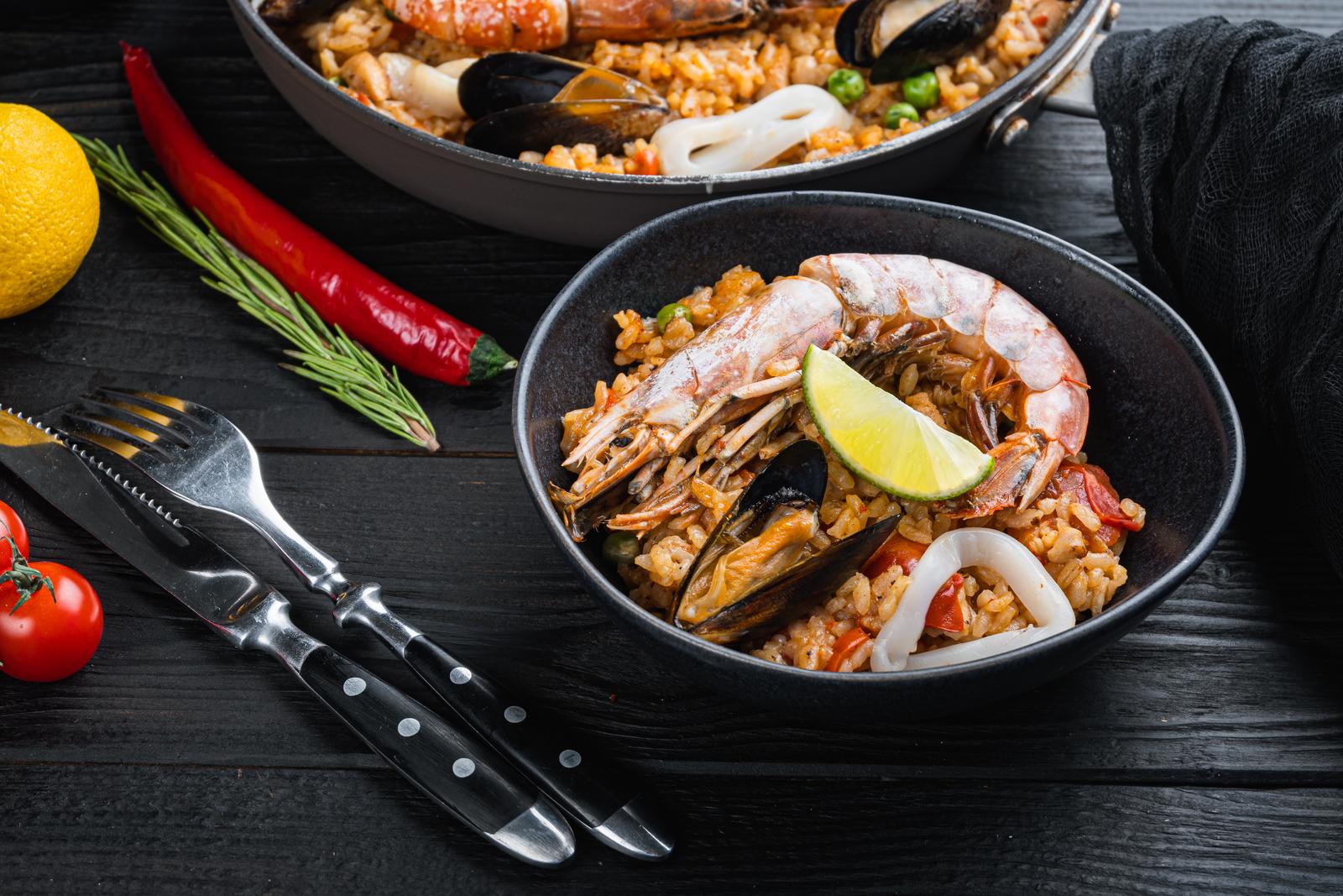 Traditional spanish seafood paella in pan and bowl with rice, peas, shrimps, mussels and squid on black wooden planks Traditional Spanish Seafood Paella In Pan And Bowl With Rice, Peas, Shrimps, Mussels And Squid On Black Wooden Planks